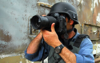 Een close-up van een persfotograaf met een helm op, gefocust door de lens van zijn camera, symboliseert de intensiteit en het belang van journalistieke fotografie.