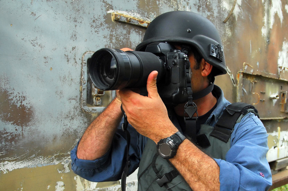 Een close-up van een persfotograaf met een helm op, gefocust door de lens van zijn camera, symboliseert de intensiteit en het belang van journalistieke fotografie.