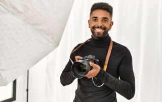 Een studiofotograaf staat in de studio met een brede glimlach op zijn gezicht en houdt een camera in zijn hand vast, wat de vreugde en enthousiasme van de fotograaf voor zijn werk in de studio uitdrukt.