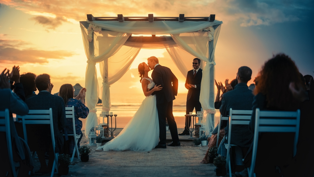 Een trouwfoto die gemaakt is op een prachtige locatie aan het strand. Op de foto staan een man en een vrouw die elkaar een kus geven voor hun familie en vrienden.