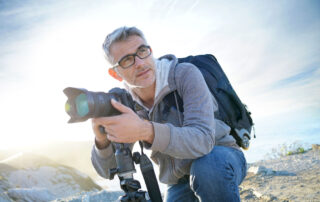 Een natuurfotograaf zit op zijn hurken met een camera in de hand, aandachtig kijkend naar de natuurlijke omgeving voor het perfecte moment om vast te leggen.