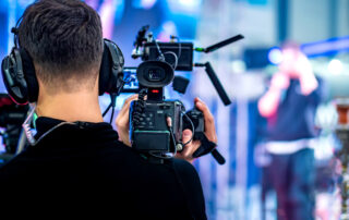 Een cameraman wordt gefotografeerd vanaf de achterkant. Het beeld toont het perspectief vanaf de achterkant van de cameraman, waarbij de camera op zijn schouder rust en gericht is op het onderwerp van de opname. Deze weergave biedt een blik op de apparatuur en de compositie zoals gezien vanuit het oogpunt van de cameraman, en benadrukt zijn rol bij het vastleggen van beelden en video's.