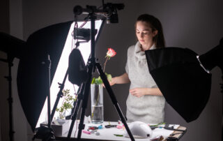 Een fotograaf gebruikt een softbox om een foto van een roos te maken in een donkere omgeving. De softbox zorgt voor een zachte en evenwichtige belichting van de roos, waardoor de details en kleuren prachtig worden vastgelegd, zelfs in het donker. Dit creëert een betoverende en sfeervolle foto.