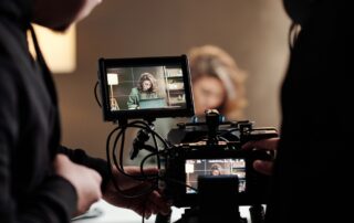 Een videograaf staat achter zijn camera en kijkt aandachtig door het scherm naar het model voor de opname. Zijn ogen zijn gefocust op het beeld dat wordt vastgelegd, terwijl hij de compositie en framing beoordeelt. De camera is gemonteerd op een statief voor stabiliteit. De scene straalt professionaliteit uit, met de videograaf in de regiestoel, klaar om het perfecte shot vast te leggen.