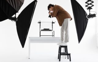 De man staat op een krukje terwijl hij een foto maakt van een product met twee verlichtingsapparaten achter hem. Dit geeft aan dat de belichting zorgvuldig is gepland om het product op de gewenste manier te fotograferen.