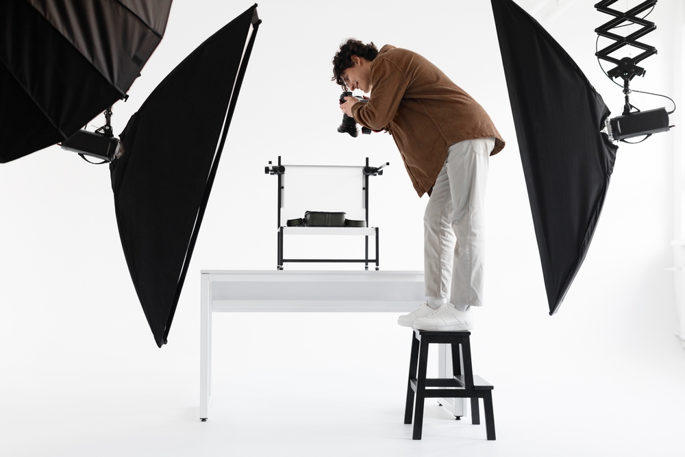 De man staat op een krukje terwijl hij een foto maakt van een product met twee verlichtingsapparaten achter hem. Dit geeft aan dat de belichting zorgvuldig is gepland om het product op de gewenste manier te fotograferen.