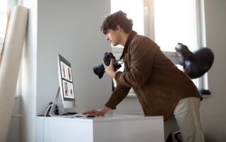 Een thuisfotostudio waarin een fotograaf staat bij een bureau, kijkend naar recent genomen foto's op een computerscherm. De fotograaf, geconcentreerd en bedachtzaam, analyseert de beelden die op het grote scherm worden weergegeven. Het bureau is uitgerust met diverse fotografie-gerelateerde voorwerpen, waaronder een camera, verschillende lenzen en mogelijk wat persoonlijke items. De omgeving suggereert een creatieve werkruimte, met misschien wat foto's of kunst aan de muur. De algemene sfeer is professioneel, maar ook comfortabel en huiselijk