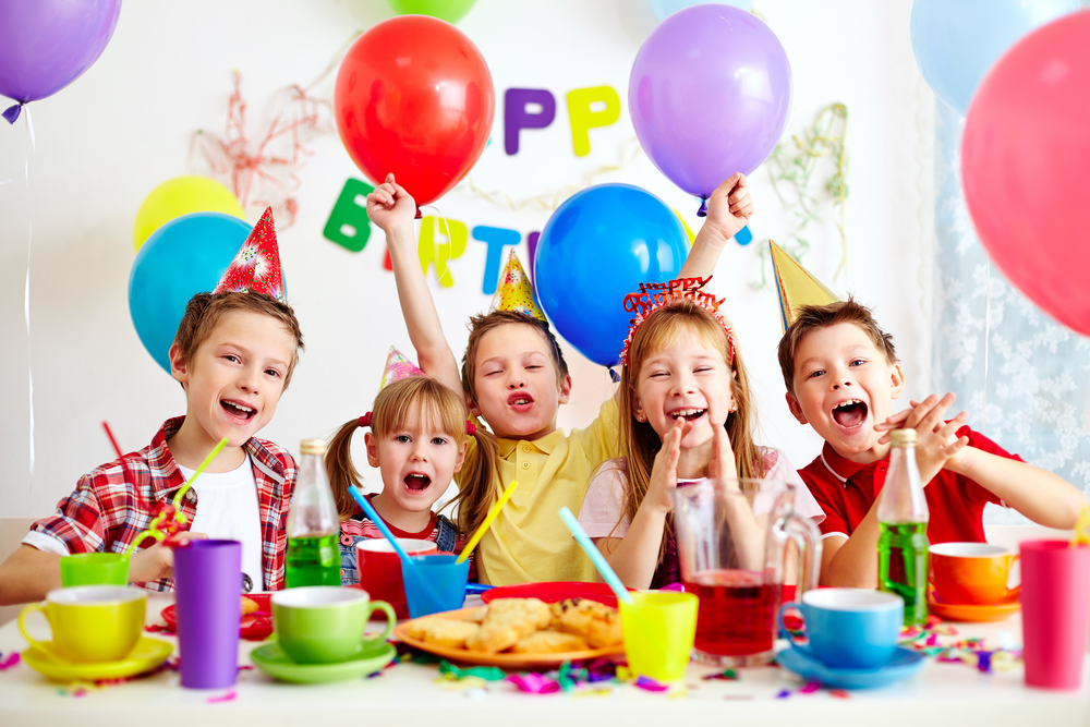 Kinderen met feesthoedjes en hun handen omhoog in een zee van licht tijdens een fotoshoot op een kinderfeestje, uitdrukking gevend aan de vreugde en feeststemming.