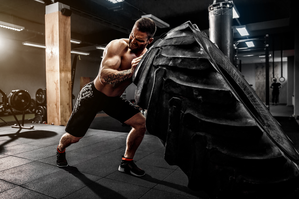 Gespierde man tijdens een fitness fotoshoot die een grote band van een vrachtwagen omduwt, demonstrerend kracht en doorzettingsvermogen in een intensieve workout setting.