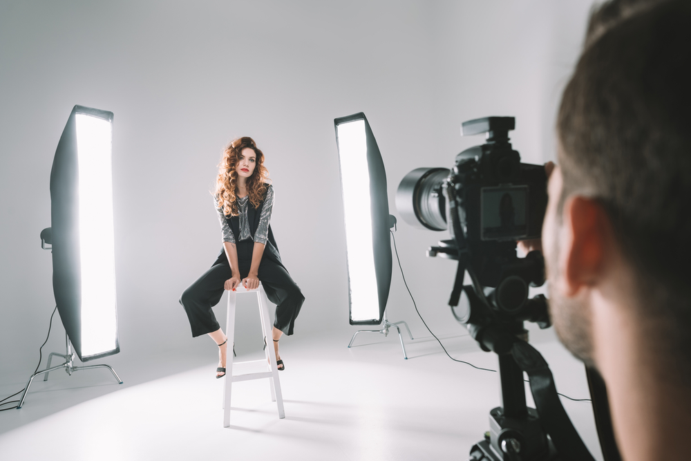 Model op een kruk met lichten en een cameraman in een studio tijdens een fotoshoot, waarbij de setting gericht is op de professionele belichting en het vastleggen van beelden.
